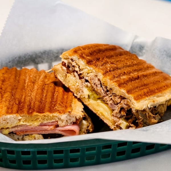 Two halves of a pressed Cuban sandwich with ham, pork, cheese, and pickles in a green basket.