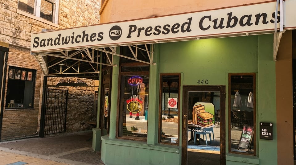 Exterior of a green building with a "Sandwiches Pressed Cubans" sign and an "Open" neon sign.
