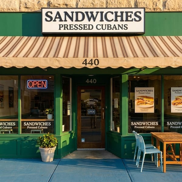 Exterior of a sandwich shop with a green facade, striped awning, and outdoor seating.